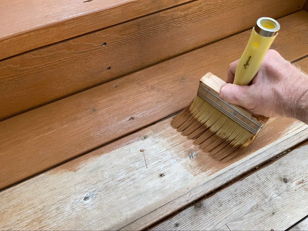 A hand uses a wide brush to apply wood stain or finish to outdoor wooden steps, covering a weathered area next to already stained wood.